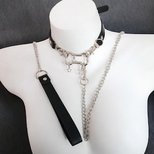 Peut inclure: Ras-de-cou en cuir noir avec un pendentif argenté en forme d'os, des breloques étoiles et une laisse assortie. Le ras-de-cou et la laisse sont reliés par une chaîne argentée. Articles présentés sur un mannequin blanc.