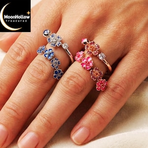 May include: Two rings featuring floral designs. One ring has blue enamel flowers with a silver-tone band and a clear stone. The other ring has pink enamel flowers with a rose gold-tone band and a clear stone. Both rings have multiple flower motifs.