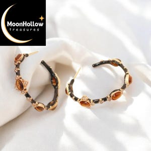 Könnte beinhalten: Ein Paar Creolen mit einem schwarz-goldenen Design. Die Ohrringe haben braune Edelsteine und kleine klare Kristalle. Der Markenname "MoonHollow treasures" befindet sich in der oberen linken Ecke.
