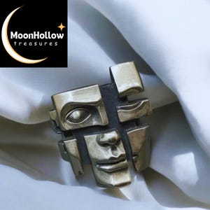May include: A silver-toned ring with a fragmented face design. The ring features a stylized human face broken into geometric shapes, with an eye, nose, and mouth visible. The ring is likely made of metal and has a vintage or artistic aesthetic. The MoonHollow treasures logo is in the upper left corner.