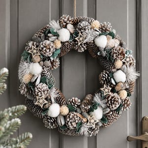 May include: A circular wreath constructed from natural brown pinecones, white cotton bolls, and small beige pom-poms. The arrangement is accented with green leaves, frosted pine needles, and several light-coloured wooden star-shaped ornaments.