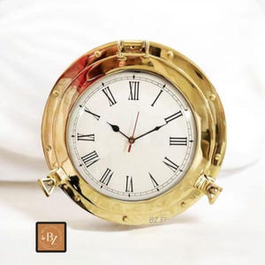 Könnte beinhalten: Eine runde, messingfarbene Uhr mit weißem Zifferblatt und schwarzen römischen Ziffern. Die Uhr hat ein nautisches Bullauge-Design mit einem Klappdeckel und seitlichen Verschlüssen. Die Zeiger sind schwarz, mit einem roten Sekundenzeiger. Das Zifferblatt zeigt "Brass Zone Elegant".