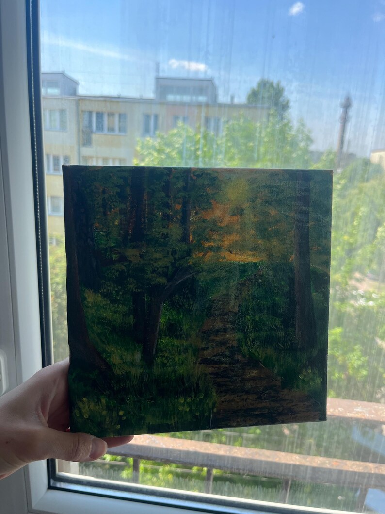 Op de afbeelding: Een vierkant canvas schilderij met een bossc&egrave;ne en een zandpad dat door donkergroene bomen slingert. Het kunstwerk toont een heldere gele en oranje gloed die door het bladerdak filtert, wat zonlicht suggereert, met diepe schaduwen en texturen.