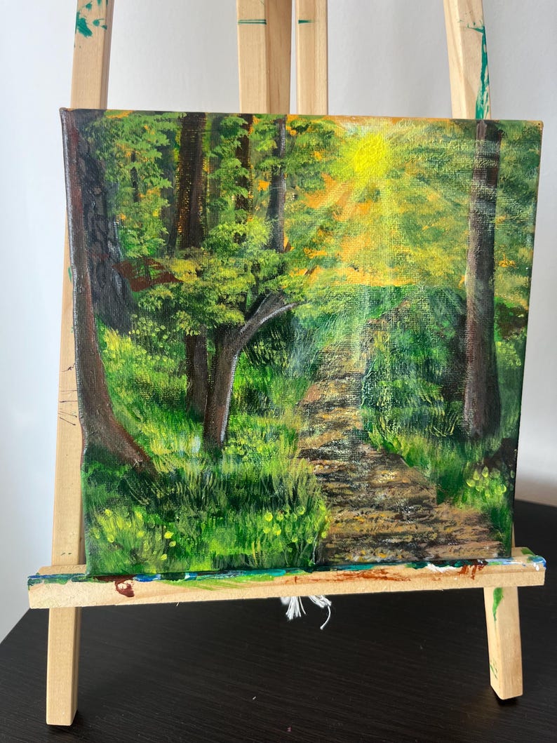 Op de afbeelding: Een vierkant canvas schilderij met een bossc&egrave;ne met een zandpad dat door groene bomen en gebladerte slingert. Een heldere gele zon schijnt door het bladerdak en werpt licht op het pad en de omliggende vegetatie in tinten groen en bruin.