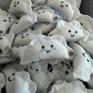 Puede incluir: Un montón de llaveros blancos con forma de nube con caras sonrientes y llaveros plateados. Los llaveros están hechos de un material suave y afelpado y tienen detalles bordados en negro. El tema general es caprichoso y lindo.