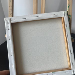 Op de afbeelding: Een vierkant opgespannen canvas op een houten frame, gezien vanaf de achterkant. Het witte doek is met metalen nietjes aan de lichtgekleurde houten latten bevestigd. De letters KT zijn met zwarte inkt op de onderste lat geschreven.