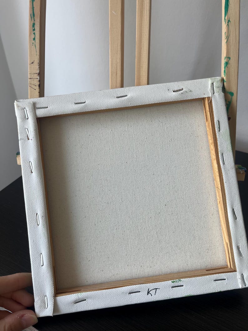 Op de afbeelding: Een vierkant opgespannen canvas op een houten frame, gezien vanaf de achterkant. Het witte doek is met metalen nietjes aan de lichtgekleurde houten latten bevestigd. De letters KT zijn met zwarte inkt op de onderste lat geschreven.