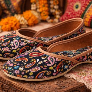 May include: A pair of black slip-on shoes with a colorful paisley pattern. The shoes have a tan trim and a light-colored sole. The shoes are displayed on a wooden surface with decorative elements in the background.