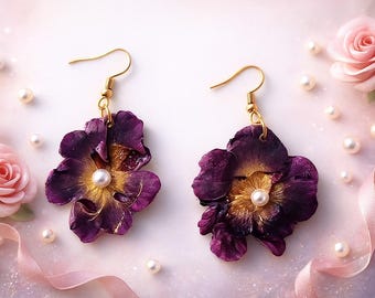 Könnte beinhalten: Ein Paar Blumenohrringe mit goldfarbenen Haken. Jeder Ohrring hat eine tiefviolette Blume mit goldenem Zentrum und einer Perle. Der Hintergrund umfasst rosa Rosen, Perlen und ein rosa Band.
