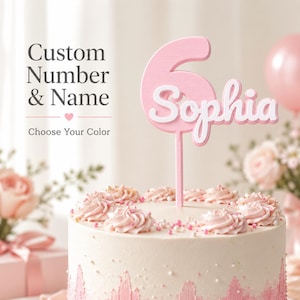 May include: A pink cake topper featuring the number 6 and the name Sophia in white script lettering. The topper is inserted into a round cake decorated with pink frosting swirls and sprinkles. Text on the image reads Custom Number & Name.