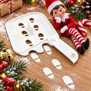 May include: A white plastic stencil tool designed for creating miniature footprint patterns with flour or powdered sugar. The stencil features a series of small oval cutouts and a handle, accompanied by a small elf doll in a red and white outfit.