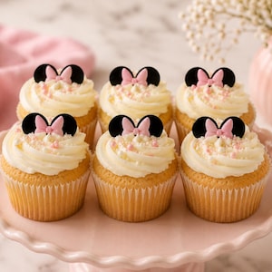 May include: Six golden-brown cupcakes topped with white frosting, small pink sprinkles, and a black and pink bow-shaped topper. The cupcakes are arranged on a light pink scalloped cake stand, featuring a design inspired by a mouse character.