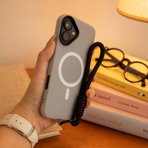 May include: A light gray phone case with a white circular design and a black lanyard. The case has a dual-camera cutout and a magnetic charging symbol. The lanyard is made of a black, braided material.