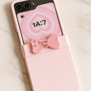 May include: A pink phone case with a bow detail. The case has a screen displaying a pink heart design and the text "1A:7". The bow features a gem in the center and cross-shaped accents on each side. The phone case is on a marble surface.
