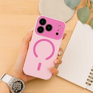 May include: A light pink phone case with a magenta camera section and a matching magenta circular design. The case has a smooth finish and is designed to fit a smartphone. The case is shown on a light wooden surface.