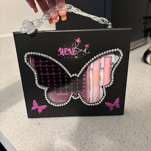 Könnte beinhalten: Schwarze Box mit einem schmetterlingsförmigen Ausschnitt, die Schönheitsprodukte enthält. Die Box hat einen transparenten Griff und ist mit Strasssteinen und rosa Schmetterlingsgrafiken verziert. Der Text "REAL Bad LASHES" ist sichtbar.