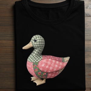 Peut inclure: T-shirt noir avec un motif de canard en tissu. Le canard a une tête à carreaux verts et blancs, un corps à carreaux roses et blancs et des motifs floraux. Le bec du canard est beige et les pieds sont beiges.