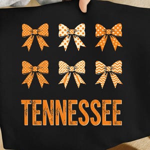 Peut inclure: T-shirt noir avec six nœuds orange à motifs variés, dont des pois et des chevrons. Le mot "TENNESSEE" est imprimé en lettres orange vieilli sous les nœuds.