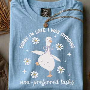 Könnte beinhalten: Hellblaues T-Shirt mit einem Cartoon-Gans-Design, Gänseblümchen und dem Text "SORRY I'M LATE. I WAS AVOIDING non-preferred tasks." Eine goldene Halskette mit Anhänger liegt auf dem gefalteten Shirt.