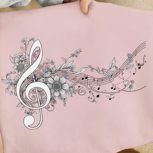 Peut inclure: Un coussin rose clair avec un motif musical noir et blanc. Une grande clé de sol est entourée d'éléments floraux et de notes de musique. Le design est un dessin au trait détaillé, créant une esthétique délicate et artistique.