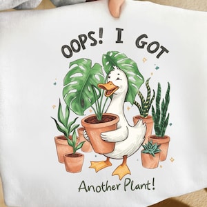 Peut inclure: T-shirt blanc avec un dessin d'un canard entouré de plantes en pot. Le canard tient une plante dans un pot en terre cuite. Le texte dit "OOPS! I GOT Another Plant!"
