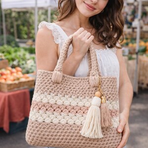 May include: A beige crochet tote bag with horizontal stripes in white and pink. The bag has two handles and a decorative tassel with a wooden bead. The bag is held by a person in a white top and blue jeans.