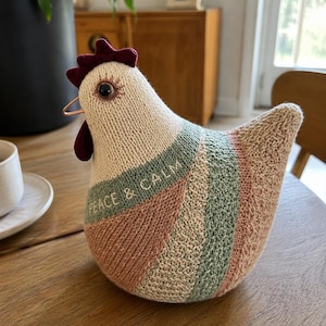 May include: A knitted chicken figurine featuring a cream-colored body with diagonal stripes in muted pink and sage green. The chicken has a dark maroon comb and wattle, a single dark eye, and the text PEACE & CALM embroidered on its side.