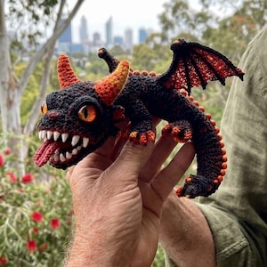 May include: A crocheted dragon figurine featuring a black body with vibrant orange accents on the horns, wings, and tail. The creature has large orange eyes, a set of sharp white teeth, and a protruding red tongue, crafted with detailed yarn texture.