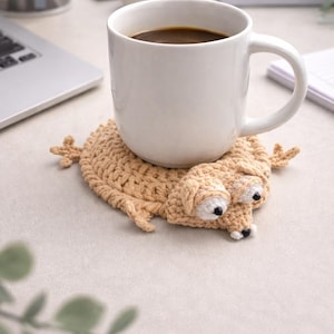 以下が含まれることがあります： 濃いコーヒーが入った白いセラミックマグが、ベージュのかぎ針編みのトカゲ型コースターの上に置かれています。 コースターには白い目と黒のアクセントがあります。 マグとコースターは明るい色の表面にあり、背景にはラップトップと緑があります。