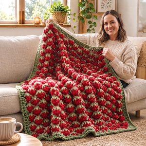 Puede incluir: Una manta tejida a ganchillo a mano con un patrón repetitivo de fresas rojas tridimensionales con hojas verdes. La manta tiene un borde grueso y texturizado en hilo verde salvia, creando un diseño de superficie denso y táctil.