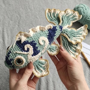 Puede incluir: Un juguete de pez koi tejido a mano con un cuerpo texturizado en tonos verde claro, azul oscuro y crema. El pez tiene ojos grandes, oscuros y brillantes con bordes dorados y aletas intrincadas con volantes ribeteadas con hilo dorado.