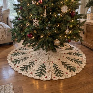 May include: A round, cream-colored crocheted Christmas tree skirt featuring a repeating pattern of dark green pine trees and small red berries. The skirt has a scalloped edge and three sets of matching cream-colored ties to secure it around the base.