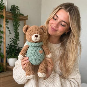 May include: A brown crocheted teddy bear with a light mint green sweater and matching scarf. The bear features a cream-colored muzzle, a dark brown embroidered nose and mouth, closed eyes, and a small heart-shaped patch on the chest.