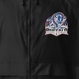 May include: A black zip-up windbreaker jacket featuring a graphic on the upper left chest. The graphic depicts a charging bison in blue, white, and red colors above a city skyline silhouette and the text BUFFALO in bold white capital letters.