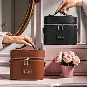 Puede incluir: Dos estuches de cosméticos personalizados, uno marrón y otro negro, con cremalleras y asas doradas. Cada estuche tiene el nombre "Lisa" en un pequeño parche rectangular. También se ve una pequeña maceta rosa con rosas rosas.