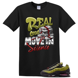 May include: A black short-sleeve t-shirt featuring a graphic print with the text Real Ones Move In Silence in gold and silver lettering, accompanied by a stack of cash. Below the shirt is a black and olive green athletic sneaker with red accents.