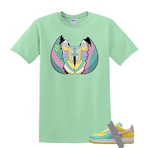 May include: A light green t-shirt featuring a graphic of sneakers inside a cracked egg. The sneakers are yellow, blue, and purple. A matching sneaker is taped to the bottom right of the shirt.
