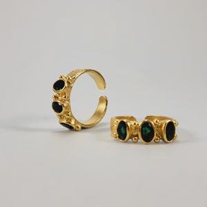 May include: Two gold-colored rings with dark green oval-shaped stones. The rings have a textured band and are open-ended, allowing for adjustable sizing. The rings are displayed on a white background.