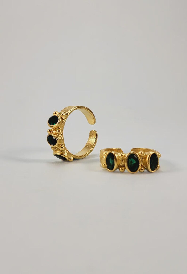May include: Two gold-colored rings with dark green oval-shaped stones. The rings have a textured band and are open-ended, allowing for adjustable sizing. The rings are displayed on a white background.