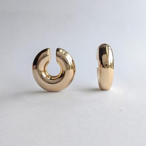 May include: A pair of polished gold-tone hoop earrings with a thick, rounded, C-shaped design. One earring is positioned to show its open circular profile, while the other is turned to display its smooth, reflective, curved outer surface.