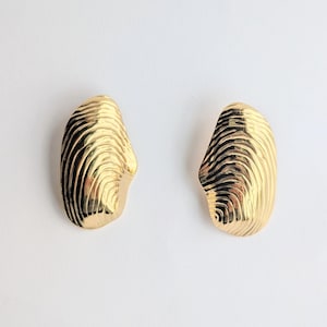 May include: A pair of gold-toned earrings with an irregular, organic shape. Each earring features a polished, reflective surface with deep, concentric, wave-like ridges that mimic the texture of a seashell or a fingerprint pattern.