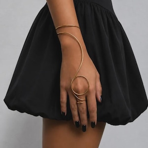 Op de afbeelding: Een goudkleurige metalen handarmband met een dun, doorlopend draadontwerp dat om de pols wikkelt en doorloopt in een cirkelvormige ring rond de middelvinger, gedragen bij een zwarte mouwloze jurk met een geplooide rok.