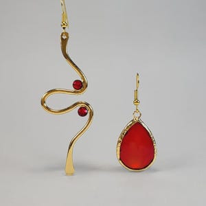 Puede incluir: Un par de pendientes dorados. Uno es un diseño estilizado y curvado con dos gemas rojas. El otro tiene forma de lágrima con una gema roja y un borde dorado. Ambos tienen cierres de gancho.