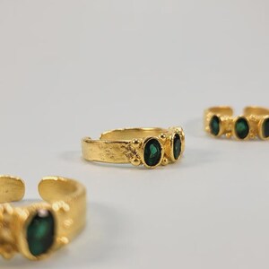 May include: Gold-toned adjustable rings with oval emerald-green gemstones. The rings have a textured band and are displayed on a white background. The gemstones are set in a row of two or three.