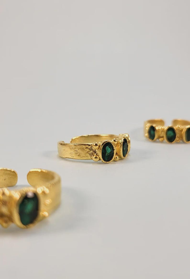 May include: Gold-toned adjustable rings with oval emerald-green gemstones. The rings have a textured band and are displayed on a white background. The gemstones are set in a row of two or three.