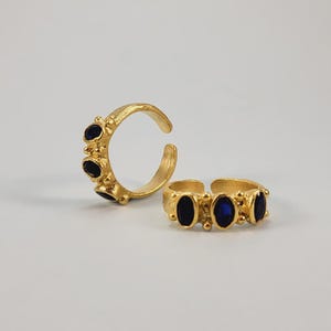 May include: Two gold-colored rings with dark blue oval-shaped stones. One ring is open and features three stones, while the other is a closed band with three stones. The rings are set against a plain white background.