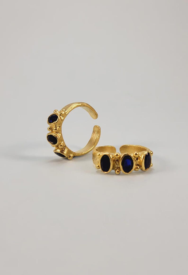 May include: Two gold-colored rings with dark blue oval-shaped stones. One ring is open and features three stones, while the other is a closed band with three stones. The rings are set against a plain white background.