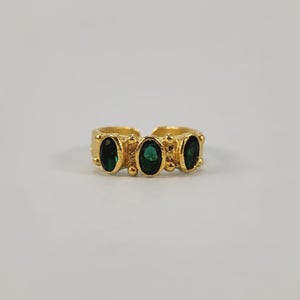 May include: A gold-toned ring featuring three oval-shaped, dark green gemstones. The ring has a textured band with small, raised details. The gemstones are evenly spaced across the top of the ring, creating a symmetrical design.