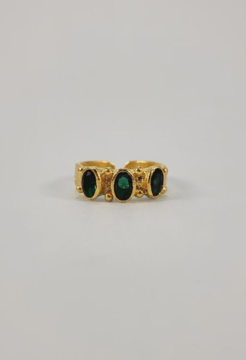 May include: A gold-toned ring featuring three oval-shaped, dark green gemstones. The ring has a textured band with small, raised details. The gemstones are evenly spaced across the top of the ring, creating a symmetrical design.