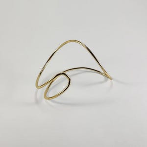 以下が含まれることがあります： ゴールドカラーの抽象的なカフブレスレット。ブレスレットは、ループと湾曲した開いた上部を備えた、連続的で流れるようなデザインが特徴です。ジュエリーは白い背景に配置され、そのミニマリストな美しさを際立たせています。